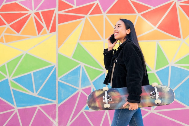 Glimlachende jonge aziatische vrouw die telefonisch praat terwijl ze loopt met een skateboard in haar hand voor een kleurrijke muur