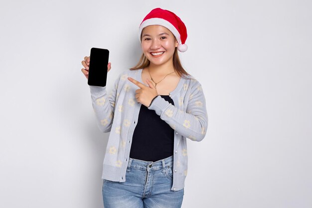 Glimlachende jonge Aziatische vrouw in een kerstmuts die een mobiele telefoon met een leeg scherm toont die mobiele app aanbeveelt die op een witte achtergrond is geïsoleerd