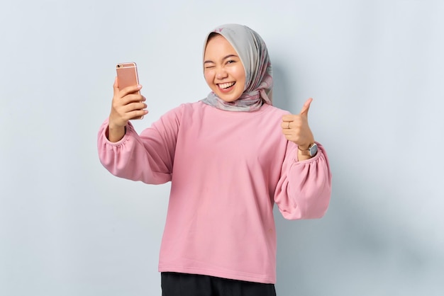 Glimlachende jonge Aziatische vrouw in roze shirt met mobiele telefoon en duim omhoog gebaar goed te keuren geïsoleerd op witte achtergrond