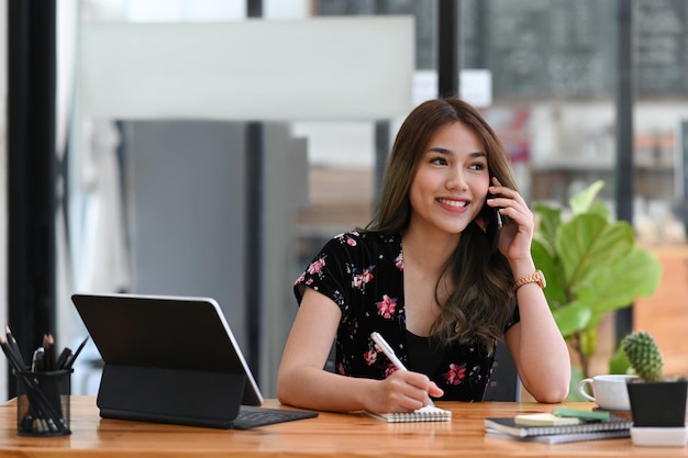 Glimlachende jonge Aziatische vrouw praten op mobiele telefoon en het maken van aantekeningen op notebook