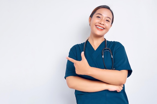 Foto glimlachende jonge aziatische vrouwelijke professionele verpleegster die een blauw uniform draagt en zelfverzekerd staat terwijl ze met de vinger wijst naar lege kopie ruimte geïsoleerd op witte achtergrond gezondheidszorg geneeskunde concept