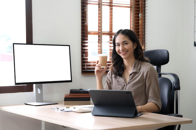 Glimlachende jonge Aziatische zakenvrouw met een koffiemok en laptop op kantoor Kijkend naar de camera