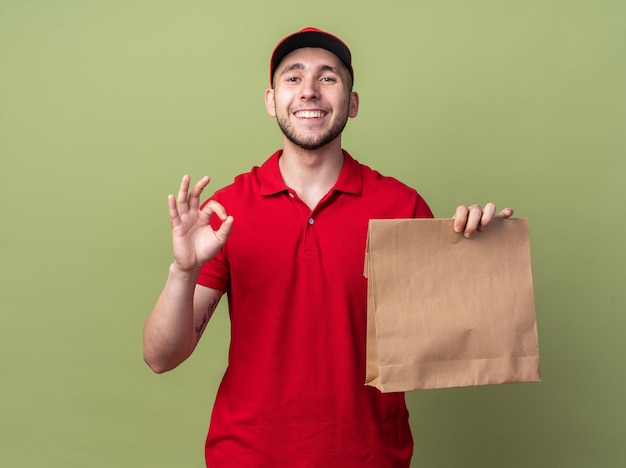 Glimlachende jonge bezorger die een uniform draagt met een pet die een papieren voedselzak vasthoudt en een goed gebaar toont