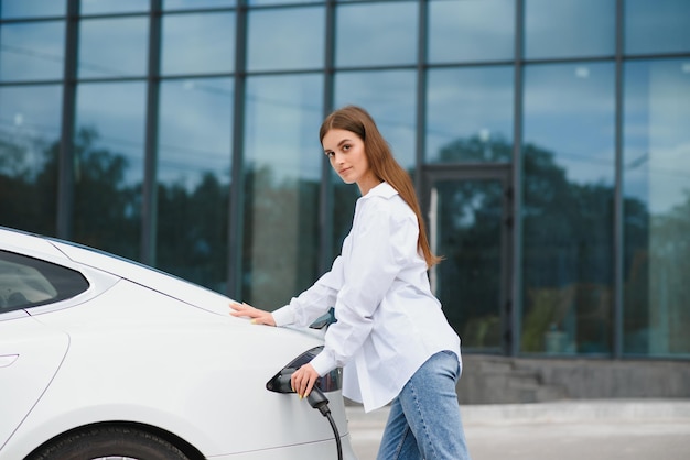 Glimlachende jonge blanke vrouw die elektriciteitskabel aansluit op elektrisch voertuig om op te laden op zonnig winkelcentrumparkeren bijgesneden lifestyle en ecologie concept