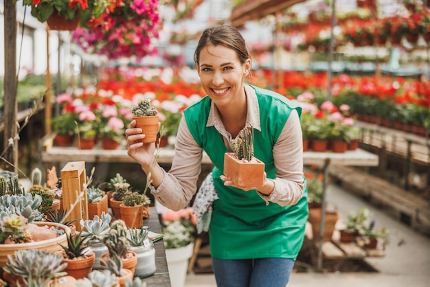 Glimlachende jonge bloemistvrouw die potten met vetplanten in een kas vasthoudt en rangschikt.