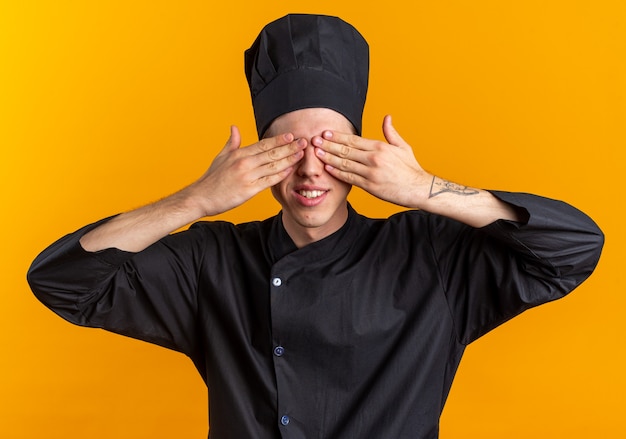 Foto glimlachende jonge blonde mannelijke kok in chef-kok uniform en pet die ogen bedekt met handen