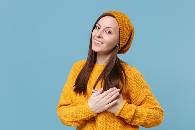 Glimlachende jonge brunette vrouw in gele trui hoed poseren geïsoleerd op blauwe muur achtergrond studio portret. Mensen emoties levensstijl concept. Bespotten kopie ruimte. Hand in hand gevouwen op borst, hart.