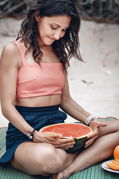 Glimlachende jonge dame houdt de helft van een watermeloen vast terwijl ze in lotushouding op het strand zit; fruit yoga