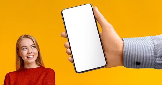 Glimlachende jonge Europese blonde vrouw die naar een enorme smartphone kijkt met een leeg scherm in de hand