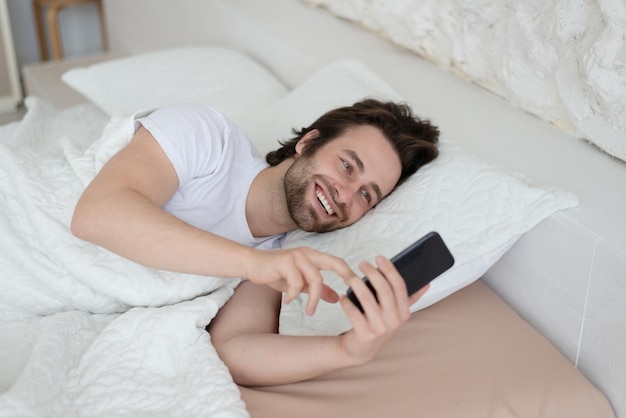 Glimlachende jonge Europese man met stoppels wordt wakker in bed terwijl hij op smartphone nieuws leest in