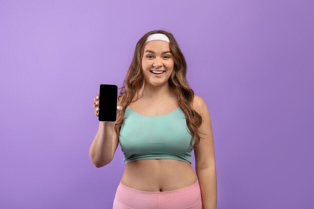 Glimlachende jonge Europese plus size vrouw in sportkleding show telefoon met leeg scherm beveelt nieuw aan