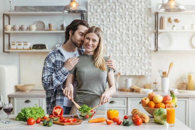 Glimlachende jonge europese vrouw bereidt salade man met stoppels knuffelt vrouw aan tafel met biologische