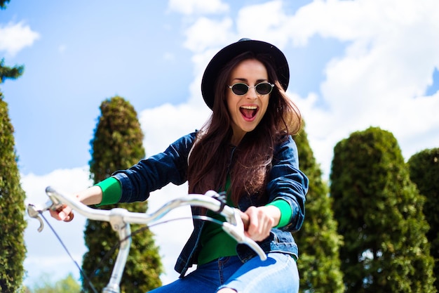 Glimlachende jonge hipstervrouw op een fiets die door de stad rijdt in een zonnebril en een hoed en plezier heeft