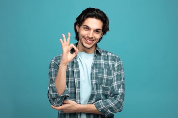 Glimlachende jonge knappe man die hand onder elleboog houdt kijkend naar camera met ok teken geïsoleerd op blauwe achtergrond