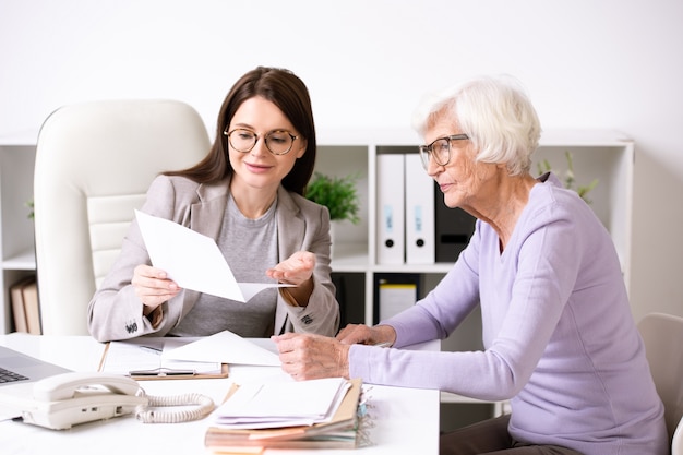 Glimlachende jonge maatschappelijk werker zittend aan tafel met senior dame en document inhoud aan haar uit te leggen