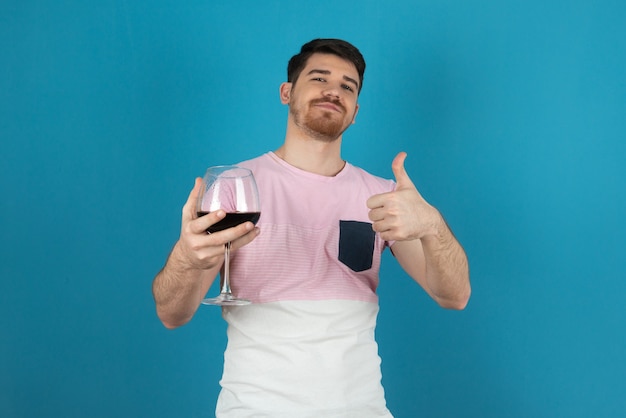 Glimlachende jonge man die een glas wijn vasthoudt en duim omhoog gebaart.