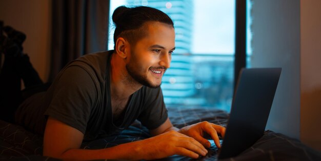 Glimlachende jonge man die op bed ligt en thuis aan zijn laptop werkt en op het toetsenbord tikt.