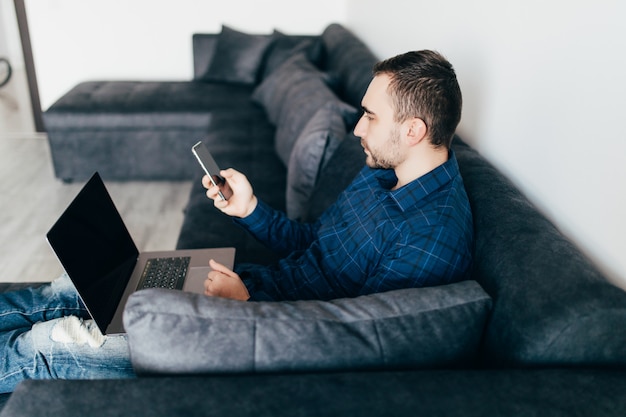 Glimlachende jonge man gebruik mobiele telefoon zittend op een bank thuis met laptop