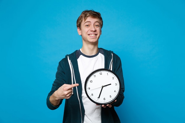 Glimlachende jonge man in casual kleding houden, wijsvinger wijzend op ronde klok geïsoleerd op blauwe muur. De tijd raakt op. Mensen oprechte emoties levensstijl concept.
