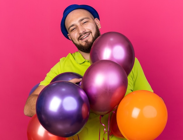 Glimlachende jonge man met feestmuts die achter ballonnen staat
