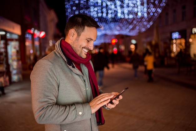 Glimlachende jonge man met mobiele telefoon op straat met Kerstdecoratie.