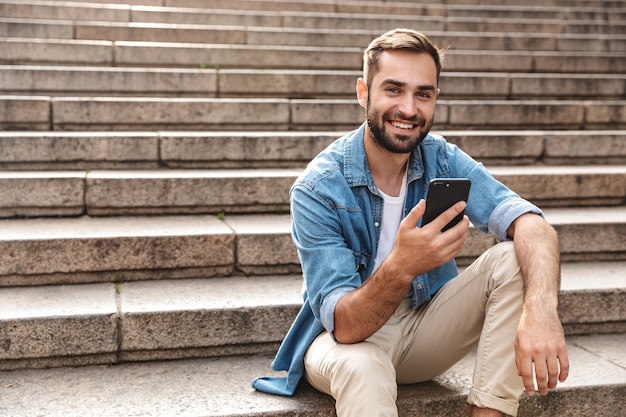 Glimlachende jonge man zittend op de trap buiten, met behulp van mobiele telefoon
