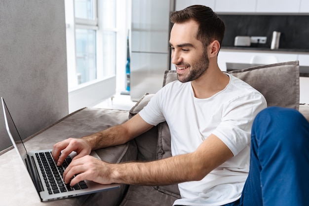 Glimlachende jonge man zittend op een bank thuis, met behulp van laptopcomputer