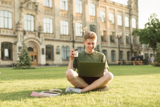 Foto glimlachende jonge man zittend op het gazon in de buurt van de universiteit met een kopje koffie in de hand en met behulp van laptop