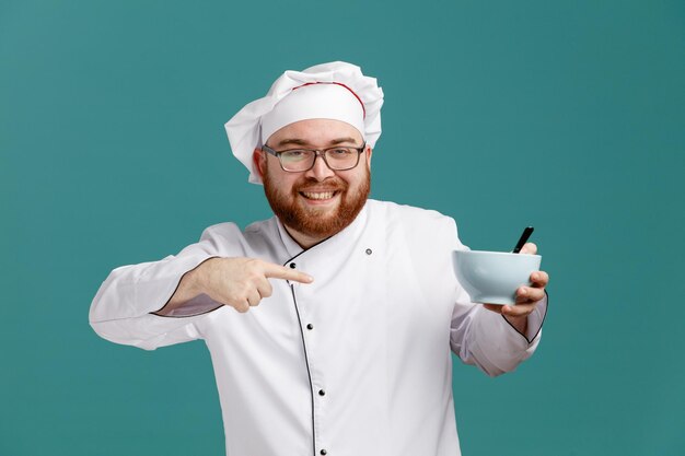 Glimlachende jonge mannelijke chef-kok met een uniforme bril en pet kijkend naar camera met lege kom met lepel erin wijzend naar kom geïsoleerd op blauwe achtergrond