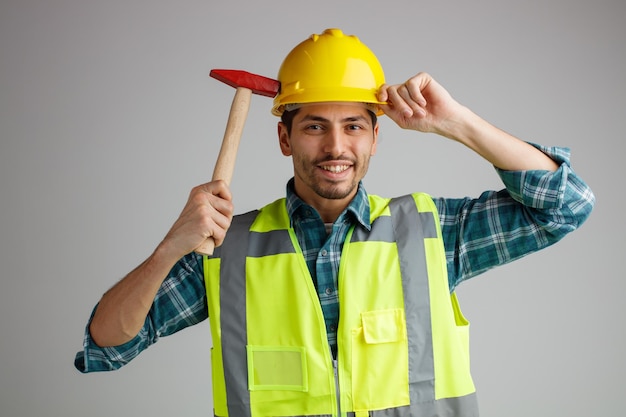 Glimlachende jonge mannelijke ingenieur met veiligheidshelm en uniform kijkend naar camera die helm aanraakt met hamer geïsoleerd op een witte achtergrond