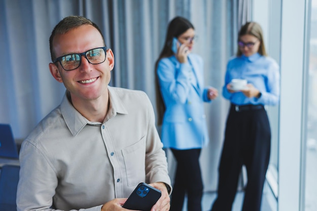 Glimlachende jonge mannelijke manager in glazen praten aan de telefoon terwijl hij op kantoor staat met collega's op de achtergrond Een werkdag in een modern kantoor met grote ramen