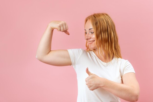 Foto glimlachende jonge roodharige vrouw met spieren op de armen portret van een mooi krachtig meisje op roze pastel achtergrond kracht van de vrouw