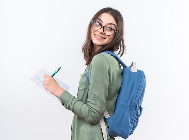 Glimlachende jonge schoolvrouw die een bril met rugzak draagt die iets op notitieboekje schrijft