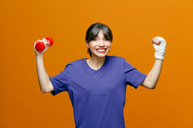 Glimlachende jonge sportieve vrouw die een t-shirt draagt met een halter die naar een camera kijkt met een sterk gebaar dat op een oranje achtergrond wordt geïsoleerd