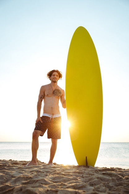 Glimlachende jonge surfer die surfplank houdt terwijl hij op het strand bij zonsondergang staat