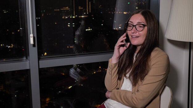 Glimlachende jonge vrouw belt en praat met een smartphone die 's nachts bij het raam zit met een panoramisch uitzicht op de stad. Brunette in glazen thuis rust na werkdag, kopieer ruimte