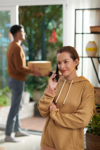 Glimlachende jonge vrouw die aan de telefoon praat wanneer haar man een geleverd pakket op de achtergrond ontvangt