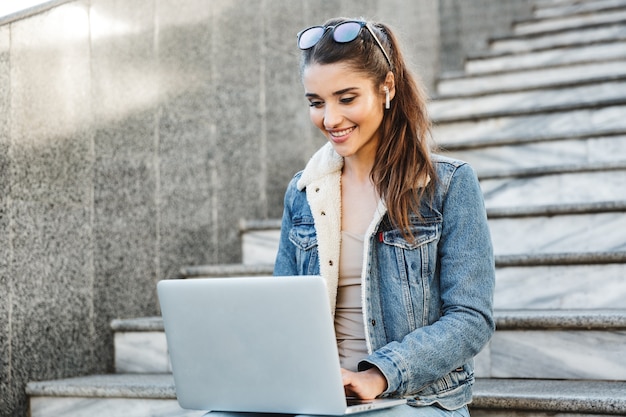 Glimlachende jonge vrouw die jas draagt die zich bij de trap in openlucht, lap top computer gebruikt