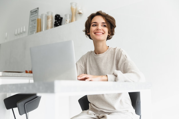 Glimlachende jonge vrouw die op laptop computer werkt zittend op een keuken