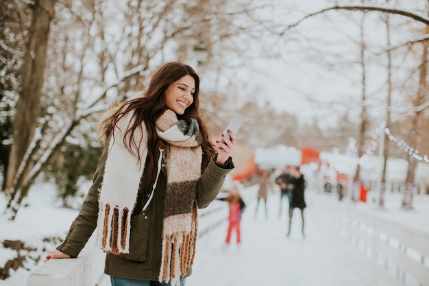 Glimlachende jonge vrouw die telefoon in park met behulp van bij koude de winterdag