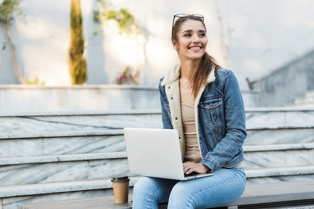 Glimlachende jonge vrouw draagt jas zittend op een bankje buitenshuis, met behulp van laptop