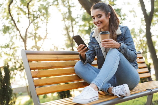 Glimlachende jonge vrouw draagt jas zittend op een bankje in het park, met behulp van mobiele telefoon, afhaalmaaltijden koffie drinken