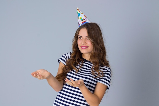 Glimlachende jonge vrouw, gekleed in gestript T-shirt en feestmuts, hand in hand voor haar op grijze achtergrond in studio