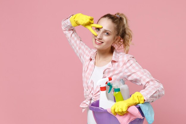 Glimlachende jonge vrouw huisvrouw in rubberen handschoenen houden bekken met wasmiddelflessen wassen reinigingsmiddelen terwijl het doen van huishoudelijk werk geïsoleerd op roze achtergrond studio. Huishoudelijk concept. Het tonen van overwinningsteken.