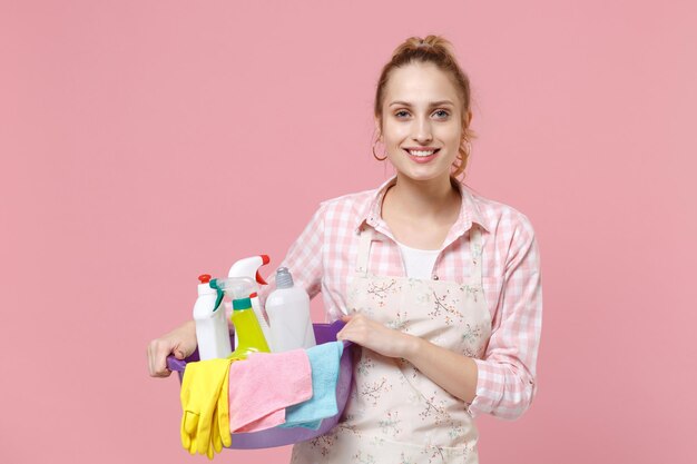 Glimlachende jonge vrouw huisvrouw in schort houden bekken met wasmiddelflessen wassen reinigingsmiddelen terwijl het doen van huishoudelijk werk geïsoleerd op roze achtergrond studio portret. Huishoudelijk concept. Camera kijken.