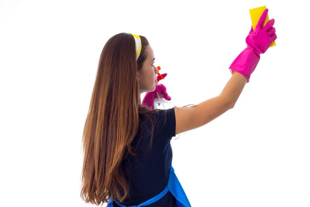 Glimlachende jonge vrouw in blauw T-shirt en schort met roze handschoenen die schoonmaakspullen in waskom houden