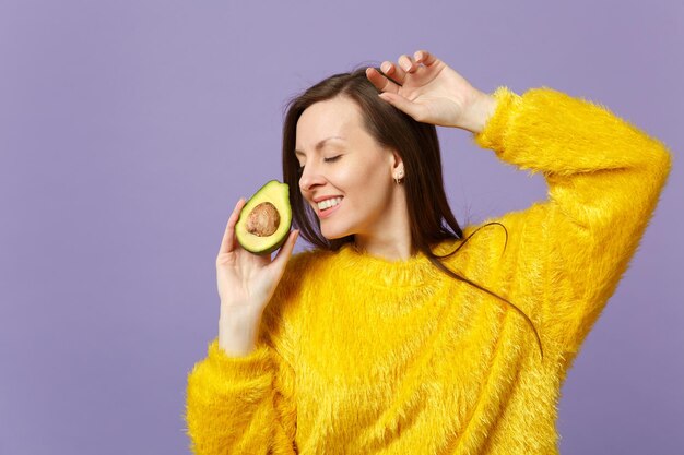 Glimlachende jonge vrouw in bonttrui die de ogen gesloten houdt met de helft van verse rijpe groene avocado geïsoleerd op violette pastelachtergrond. Mensen levendige levensstijl ontspannen vakantie concept. Bespotten kopie ruimte.