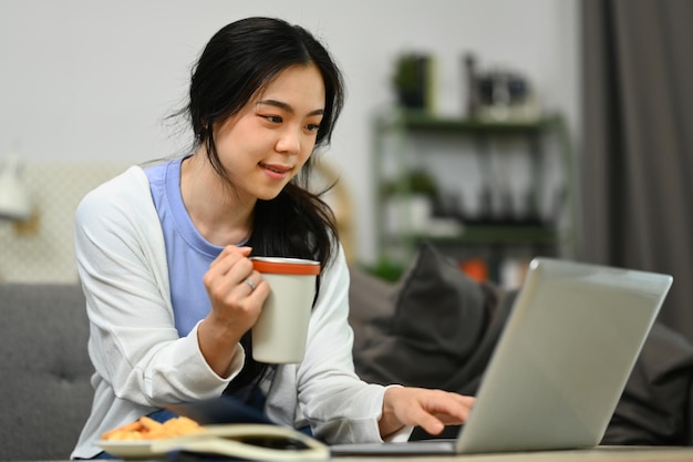 Glimlachende jonge vrouw in casual kleding met behulp van laptop op de bank thuis Mensen technologie en levensstijl