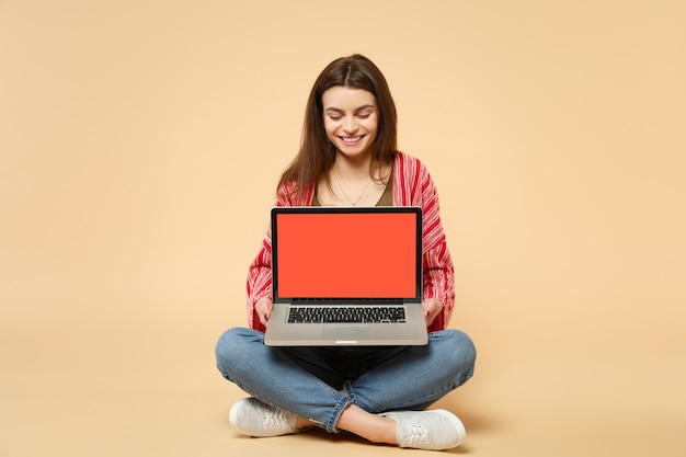 Glimlachende jonge vrouw in casual kleding zitten, met laptop pc-computer met leeg leeg scherm geïsoleerd op pastel beige achtergrond. Mensen oprechte emoties, lifestyle concept. Bespotten kopie ruimte.