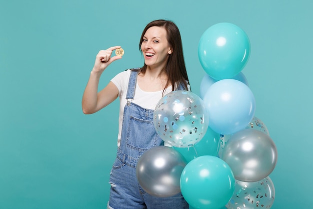 Glimlachende jonge vrouw in denim kleding met bitcoin, toekomstige valuta, vieren met kleurrijke luchtballonnen geïsoleerd op blauwe turkooizen achtergrond. Vakantie verjaardagsfeestje, mensen emoties concept.